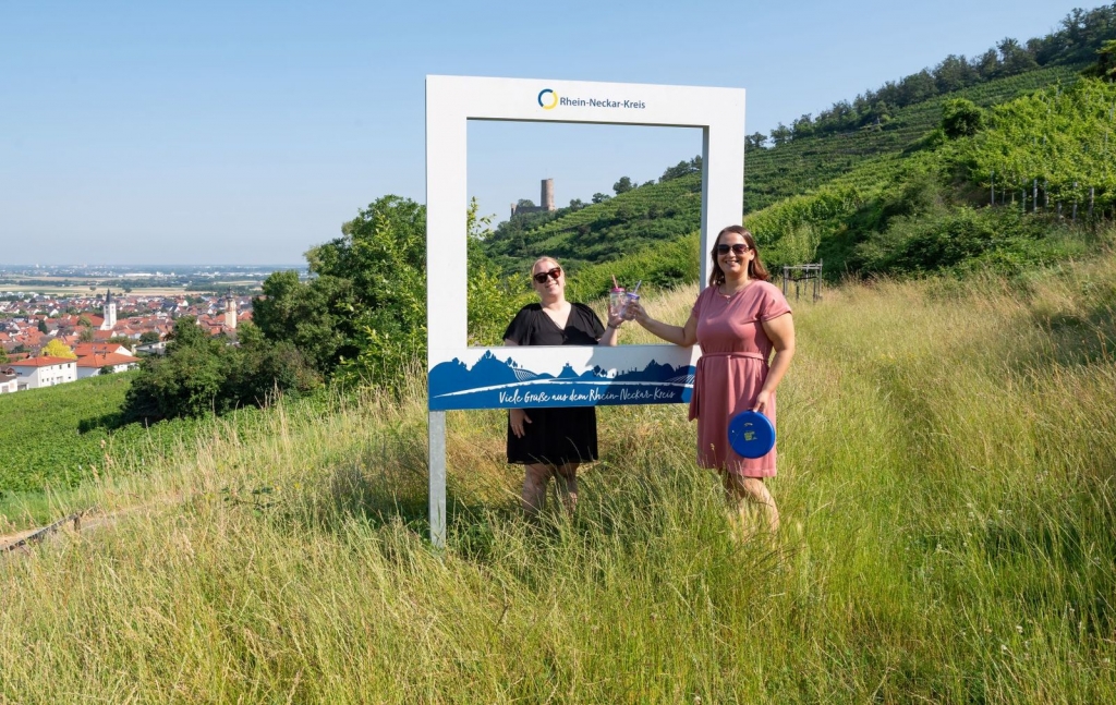 Bildunterschrift (Quelle: Landratsamt Rhein-Neckar-Kreis): Beim Fotowettbewerb „Sommer im Kreis“ sind der Kreativität keine Grenzen gesetzt – die einzige Bedingung: Das Foto muss mit einem der fünf Landschaftsfotorahmen aufgenommen werden. Hier ein Beispiel, zwei Frauen prosten sich durch den Rahmen zu, der auf einer Wiese steht.
